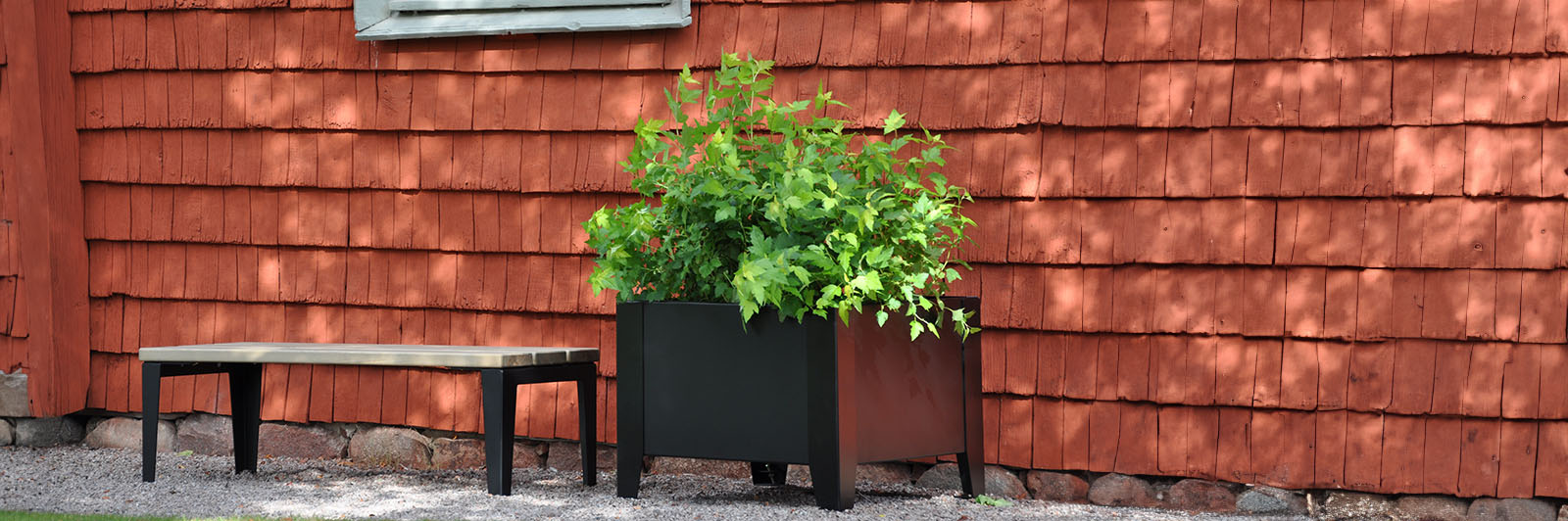 Un banc avec un grand bac à fleurs noir avec une plante verte à l'intérieur. Ils sont situés à côté d'un mur de briques dans une rue.